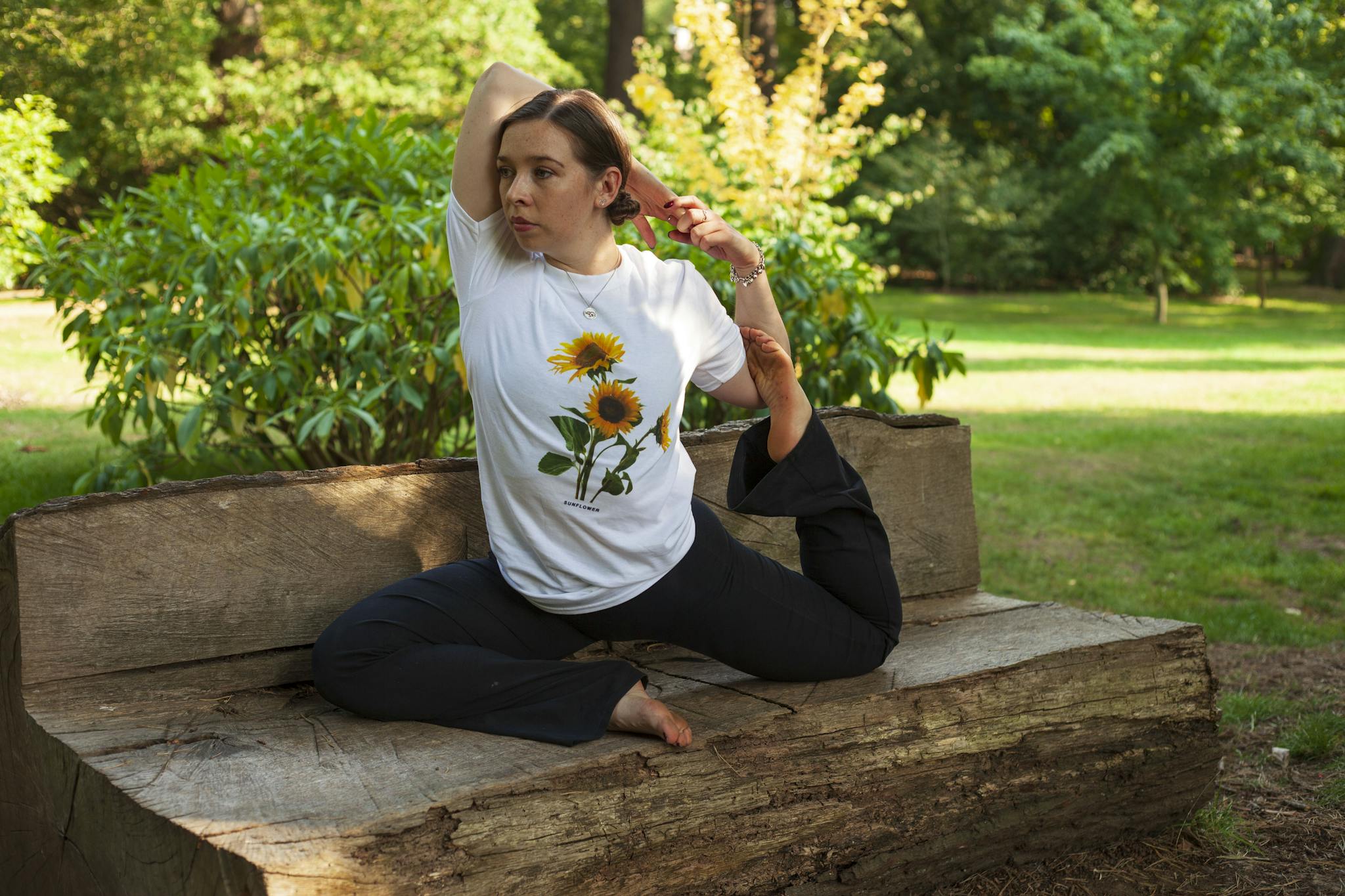About Woman performs yoga on a wooden log bench in a lush green park, promoting wellness and flexibility.
